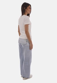 Femme debout de profil portant un t-shirt blanc uni, un jean large bleu clair avec des détails en strass, et des baskets blanches.