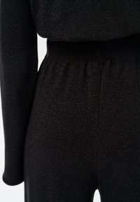 Czarny kombinezon wykonany z teksturowanej, elastycznej tkaniny o subtelnym połysku. Pas jest ściągany prążkowanym materiałem, co zapewnia dopasowaną sylwetkę.