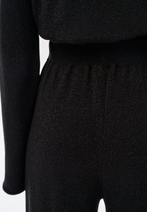 Schwarzer Overall aus strukturiertem, elastischem Stoff mit dezentem Schimmer. Der Taillenbund ist gerippt und sorgt für eine figurbetonte Silhouette.