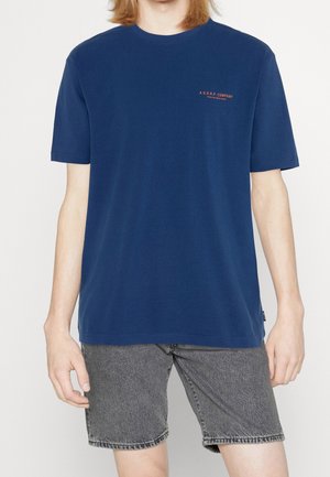 Personne portant un t-shirt bleu marine uni avec un petit texte orange sur la poitrine et un short en jean gris délavé, debout devant un fond neutre.