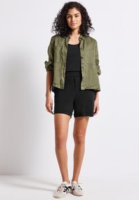 Camicia leggera verde oliva con bottoni sopra una canotta nera e pantaloni corti neri. Le sneakers casual completano l'outfit. Tessuto morbido in tutta la lunghezza.