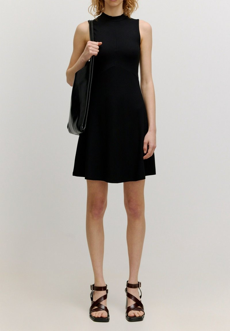 Femme portant une robe noire sans manches, tenant un sac à bandoulière noir, et portant des sandales marron à talons hauts et à brides, debout devant un fond uni.