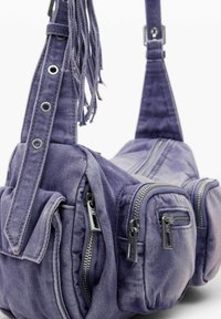 Ljubičasta platnena torba s višestrukim zatvorenim džepovima, pletenim remenom s metalnim detaljima i teksturiranom završnom obradom, koja ima resice na remenu.