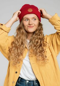 Czerwona dziana czapka z okrągłą skórzaną łatą; noszona z żółtą kurtką na białej koszuli i niebieskich dżinsach. Miękkie, teksturowane włosy.