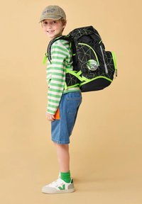 Black backpack with neon green accents and geometric design. Child wearing green striped shirt, denim shorts, and white shoes with green accents.