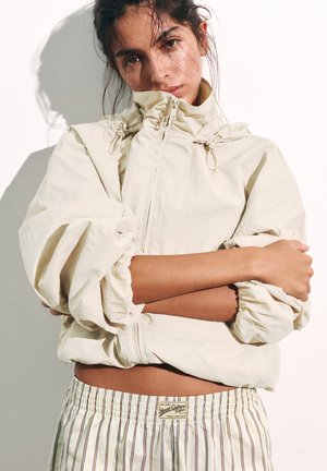 Un mannequin prend la pose, les bras croisés, vêtu d’une veste crème zippée et d’un short à rayures, devant un fond de studio uni.