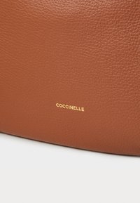 Braune Lederhandtasche mit strukturierter Oberfläche und einem goldenen, geprägten Logo "Coccinelle" auf der Vorderseite.