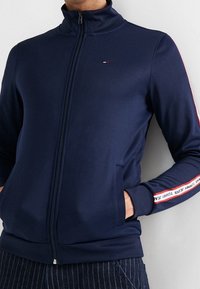 Navy blue zip-up jacket made of smooth fabric. Features red and white stripes along sleeves and a small logo on the chest. Two side pockets.