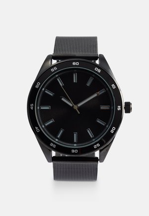 Orologio analogico nero con cassa metallica rotonda, quadrante nero, indicatori delle ore luminescenti e cinturino grigio testurizzato. Presenta un design minimalista.