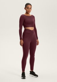 Burgundská dlhá rukávová crop top spojená s legínami s vysokým pásom, ktoré majú design prekríženého pásu, vyrobené z elastického a hladkého materiálu.