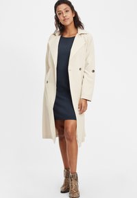 Beige trench coat suure nupud ja lai krae, kantud tumedast sinisest, keha rõhutavast kleidist. Kandub koos käärmöödud pahkluu saapatega.
