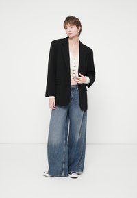 Černý oversize blejzr, krémový zkrácený svetr s knoflíky, vybledlé modré široké džíny, texturovaná tkanina, oblečené s bílými teniskami.