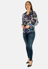 Blouse à manches longues en voile violet, vert et bleu à imprimé floral, associée à un jean skinny bleu foncé et des escarpins noirs.