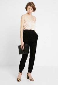 Růžový hedvábný top s knoflíky, kombinovaný s černými tepláky s krajkovým vzorem a černou taškou. Modelka má na sobě zelené podpatky a stojí na bílém pozadí.