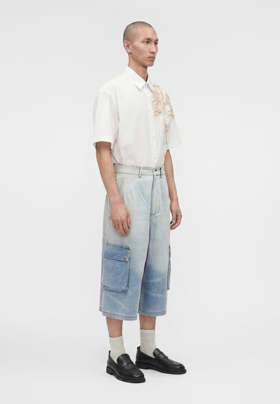 Άνδρας μοντέλο φοράει λευκό κοντομάνικο πουκάμισο με κεντήματα λουλουδιών, ανοιχτό μπλε φαρδιά τζιν σορτς με μεγάλες τσέπες, λευκές κάλτσες και μαύρες μοκασίνες.