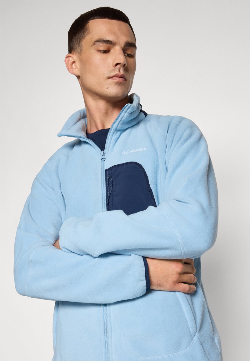 Jasnoniebieska polarowa kurtka z wysokim kołnierzem, zapinana na zamek, z ciemnoniebieską kieszenią na piersi. Miękka tekstura i luźny fason.