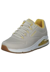 Beige-tossu, jossa perforoitu design, keltaisia yksityiskohtia, pehmustettu kaulus ja läpikuultava ilmatyynyvälipohja. Varustettu tasaisilla nauhoilla ja kumipohjalla.