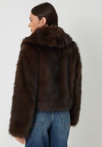 Braune Kunstfelljacke mit Kapuze, cropped Design und langen, weiten Ärmeln. Die Textur wirkt weich und dicht mit einem leichten Schimmer.