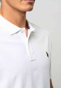 Mężczyzna w białej koszulce polo z małym ciemnym logo konia i jeźdźca na piersi oraz granatowymi czerwonymi paskami wewnątrz kołnierza.