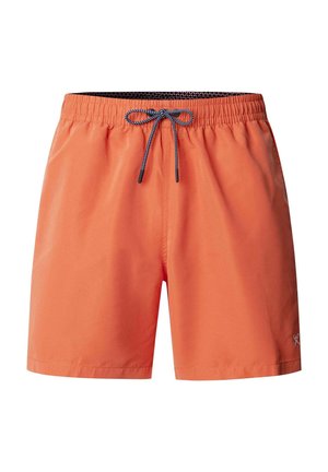 Hombre sin camiseta con pantalones cortos de baño naranja con cordón gris y zapatos marrones sin cordones, de pie contra un fondo gris liso.