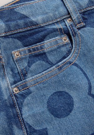 Material denim cu nuanță albastră și un model subtil de cercuri variate. Evidențiat cu cusături maro și nituri metalice pe buzunare.