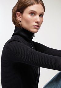 Maglione nero a coste con colletto alto e zip a metà, indossato con jeans blu chiaro. Primo piano del colletto e della spalla.