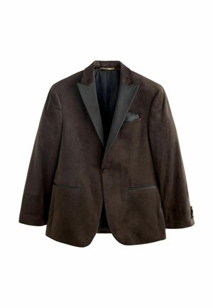 Fiatal férfi fekete blézerben, sötét ingben, bézs nadrágban és fekete alkalmi cipőben áll egy fa padlón, fehér kazettás fal előtt.