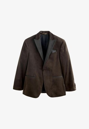 Mladý muž v čiernom blejzeri, tmavej košeli, béžových nohaviciach a čiernych spoločenských topánkach stojaci na drevenej podlahe oproti bielej stenene s panelmi.