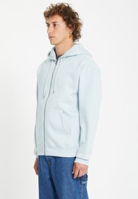 Sweat à zip bleu clair en tissu doux, doté d'une capuche avec cordon de serrage, de poches avant et de poignets à côtes, associé à un jean bleu.