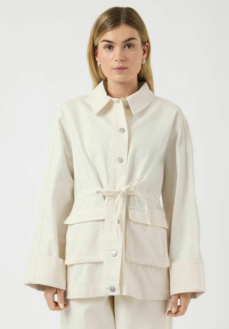 Femme portant une veste écrue boutonnée avec une ceinture à la taille et de grandes poches avant, debout devant un fond blanc uni.