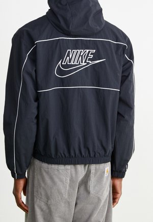 Person iført en sort Nike-jakke med hvide rør og logo på bagsiden, kombineret med lysegrå kordbukser.