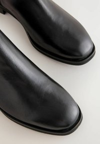 Närbild av två eleganta svarta läderstövlar, som visar deras polerade yta och rundade tår mot en ljus bakgrund.