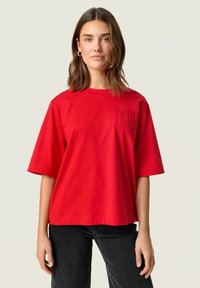 Red cotton t-shirt with a boxy shape, short sleeves, and a textured pocket detail on the left chest. Paired with black trousers.