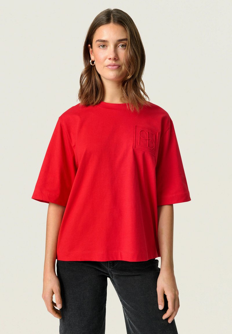 Red cotton t-shirt with a boxy shape, short sleeves, and a textured pocket detail on the left chest. Paired with black trousers.