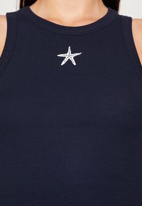 Námořnická modrá ribbed tílko s vyšitým bílým mořským hvězdicím na prsou. Tkanina je hladká, s přiléhavým střihem a bez rukávů.