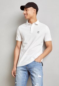 Hvid polo skjorte med krave og tre knapper, med et lille sort logo på venstre bryst. Kombineret med lys blå slidte jeans.