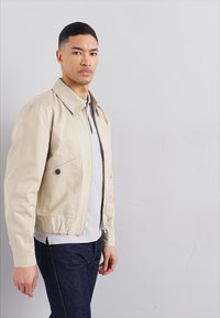 Beige műbőr dzseki, cipzáras záródással, hegyes gallérral és oldalsó zsebekkel. Karcsú szabású, minimalista designnal és elülső gomb részletekkel.