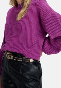 Fialový svetr z žebrovaného úpletu s vysokým límcem a širokými rukávy, kombinovaný s černými kalhotami z umělé kůže s vysokým pasem a páskem s nýty.