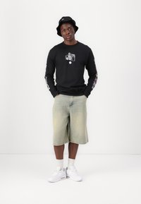 Langermet svart T-skjorte med grafisk trykk, lyse denimshorts, hvite joggesko og en svart bøttehatt, som står mot en ensfarget bakgrunn.