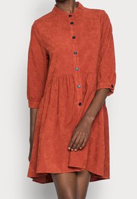 Robe couleur rouille avec col, ouverture boutonnée sur le devant, manches trois-quarts et partie inférieure évasée. Confectionnée dans un tissu texturé.