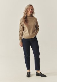 Béžový sveter s vysokým golierom, kombinovaný s tmavomodrými džínsami s priamymi nohavicami a čiernymi mokasínami, predstavený na neutrálnom pozadí.