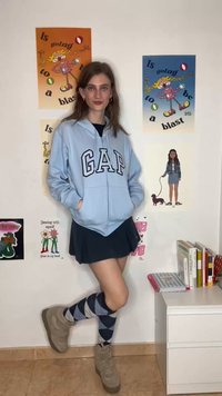 Jeune femme portant un sweat à capuche GAP bleu clair, une jupe noire, des chaussettes à losanges, des bottes beiges, debout contre un mur orné d'affiches colorées et à côté d'une commode blanche.