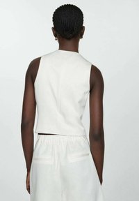 Weiße ärmellose Bluse mit hohem Halsabschluss, strukturiertem Stoff und kürzender Form, kombiniert mit lockeren, weißen Hosen mit elastischem Bund und Seitentaschen.