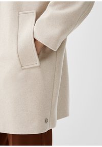 Světlý béžový kabát s texturovanou tkaninou, bočními kapsami a patkou na dolním lemu. Design má volný střih a klasický styl.