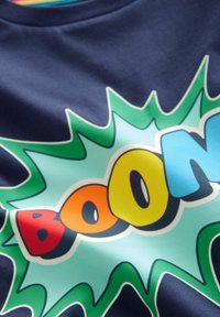 Granatowa koszulka z kolorowym graficznym napisem "BOOM" w stylu komiksu w kolorach żółtym, pomarańczowym i niebieskim, otoczona zielonymi akcentami na jasnym tle.