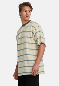 Camiseta de manga corta a rayas en color beige claro, con rayas horizontales azules, un cuello redondo marrón y un bolsillo en el lado izquierdo del pecho.