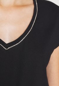 Černý pletený top s výstřihem do V, zdobený stříbrnou aplikací podél okraje. Krátké rukávy doplňují design.
