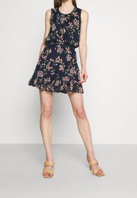 Robe marine à motifs floraux avec un design sans manches, un ourlet à volants en couches, un tissu doux et une taille cintrée. Portée avec des sandales à talons beiges.