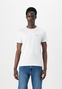 T-shirt bianca in cotone con maniche corte, colletto rotondo e logo discreto sul lato sinistro del petto. Abbinata a jeans blu in denim.