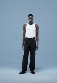 Regata branca sem mangas, jeans pretos de perna reta e sapatos pretos. Modelo está posado contra um fundo azul claro com postura estruturada.